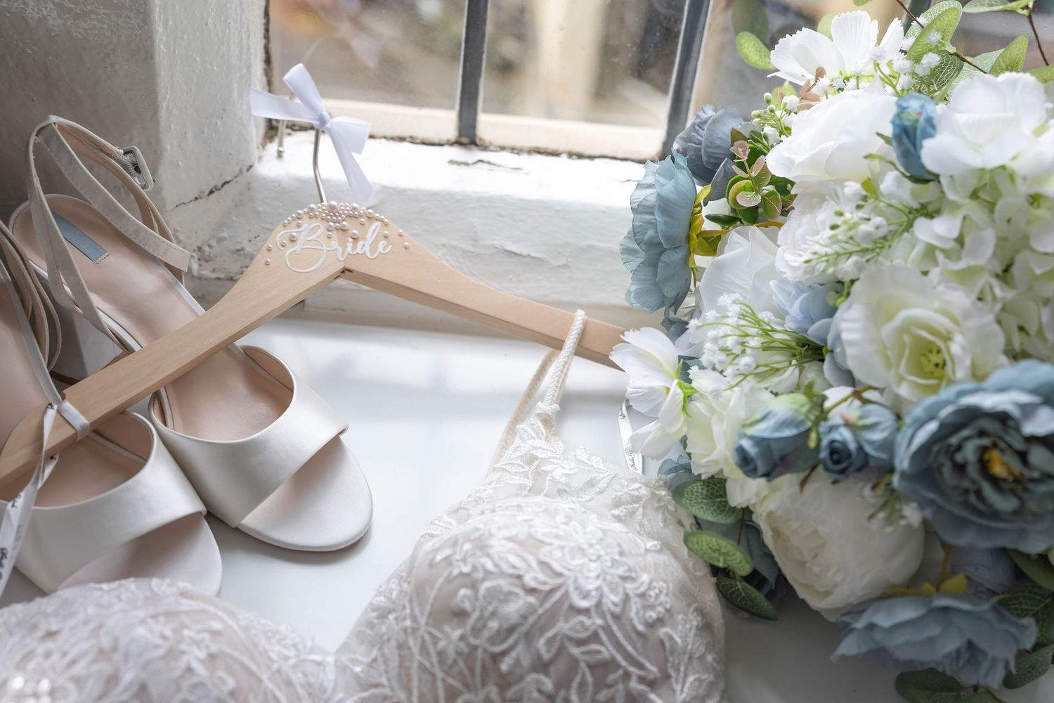 Wedding flat lay featuring bridal heels, floral bouquet with blue roses, and lace details on windowsill.