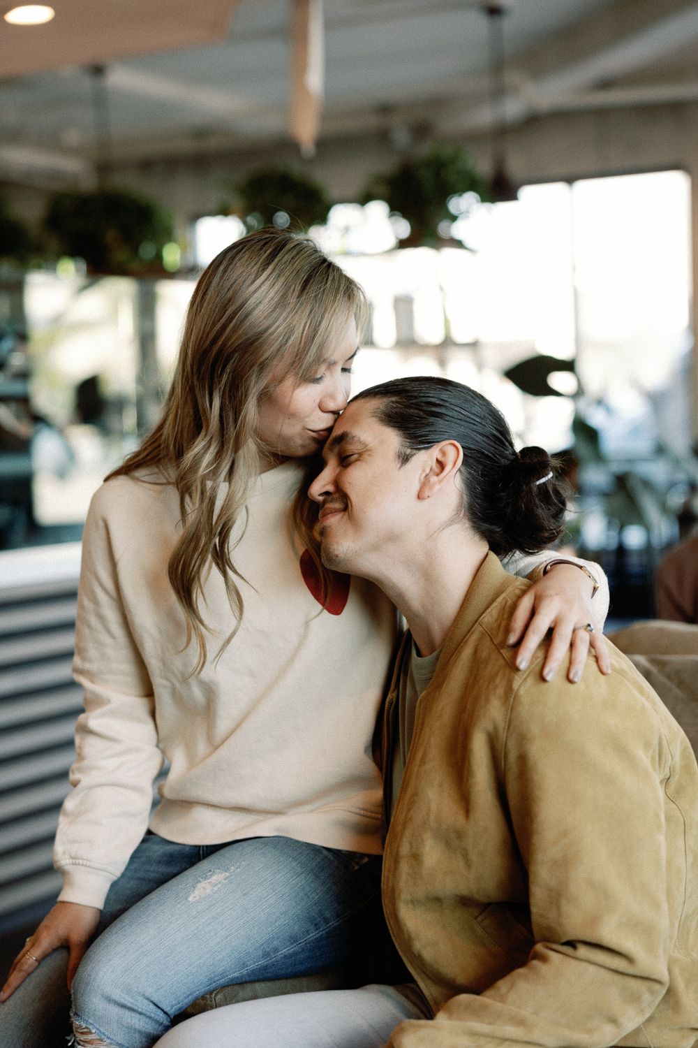 A woman kisses the forehead of a smiling man with a bun hairstyle in a cozy cafe setting.