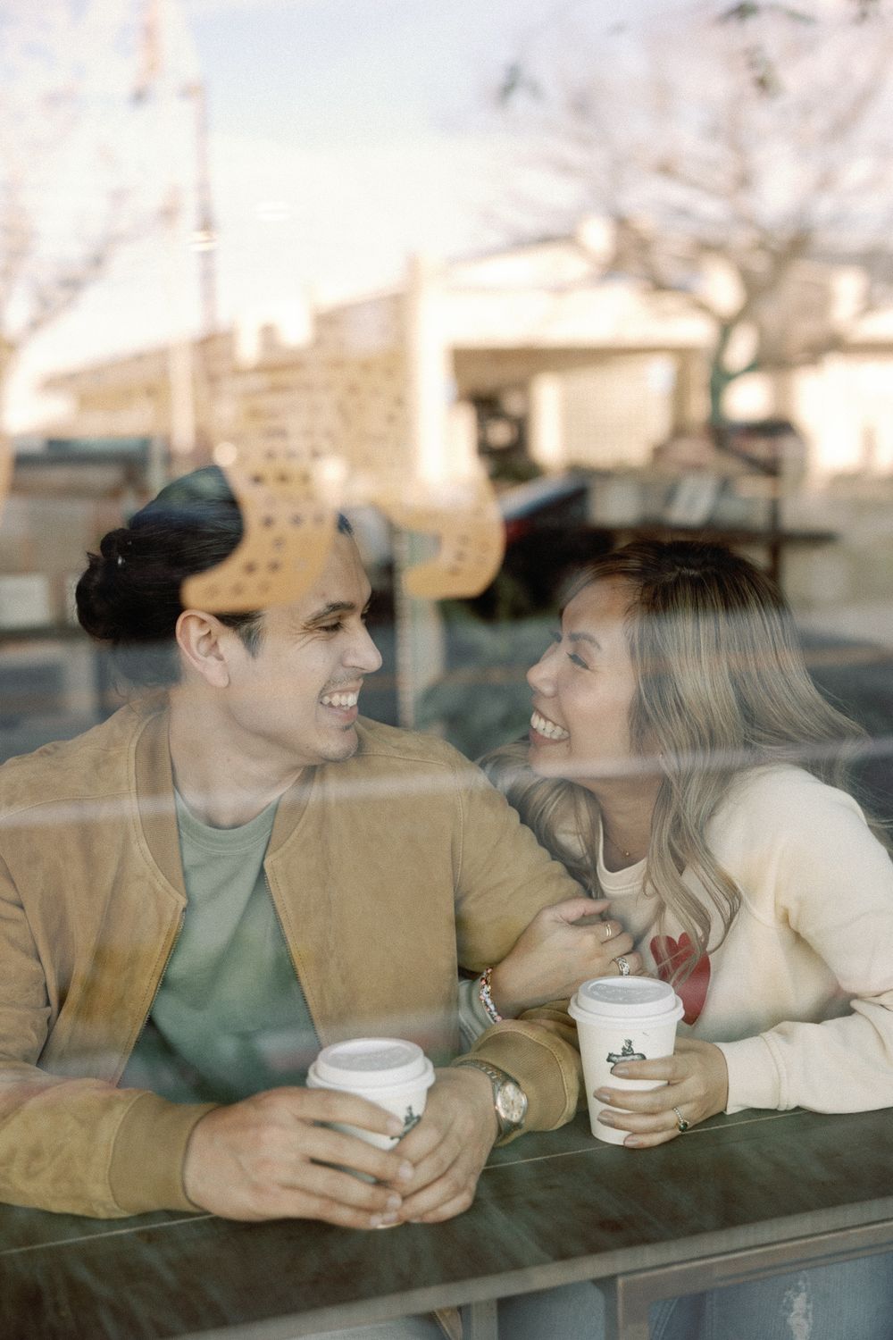 Couple smiling at each other through cafe window, holding takeaway coffee cups on a date.