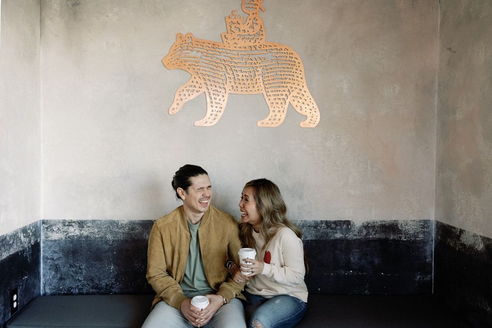 A couple smiles at each other while holding coffee cups, seated beneath an orange bear wall art.