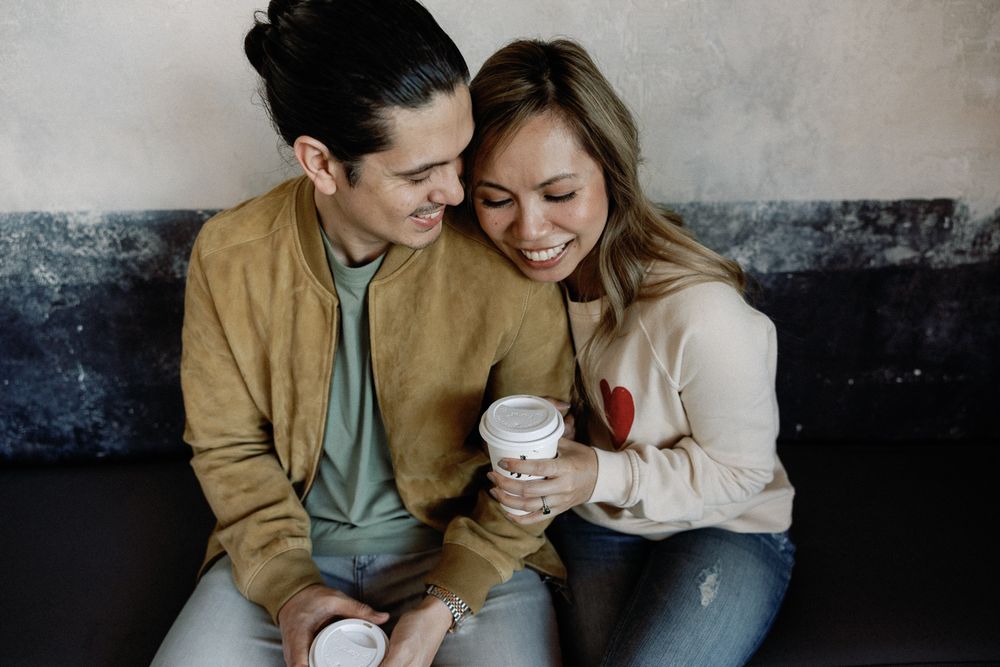 Happy couple sitting together on a couch, sharing coffee cups and smiling in a cozy cafe setting.