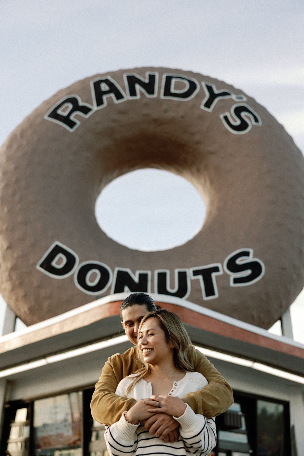 Couple embracing romantically in front of the iconic Randy's Donuts giant donut sign in Los Angeles.