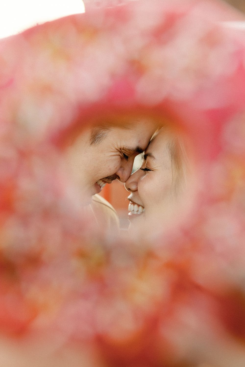 Couple smiling face-to-face framed by soft pink bokeh flowers in a romantic close-up portrait.