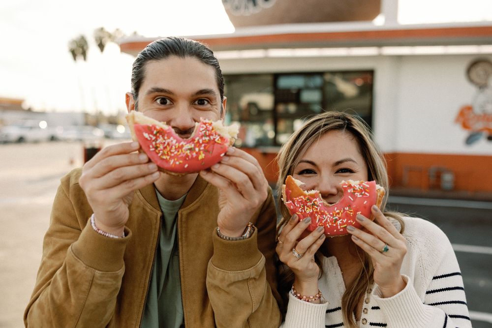 Couple holding large pink frosted sprinkle donuts up to their faces outside a donut shop.