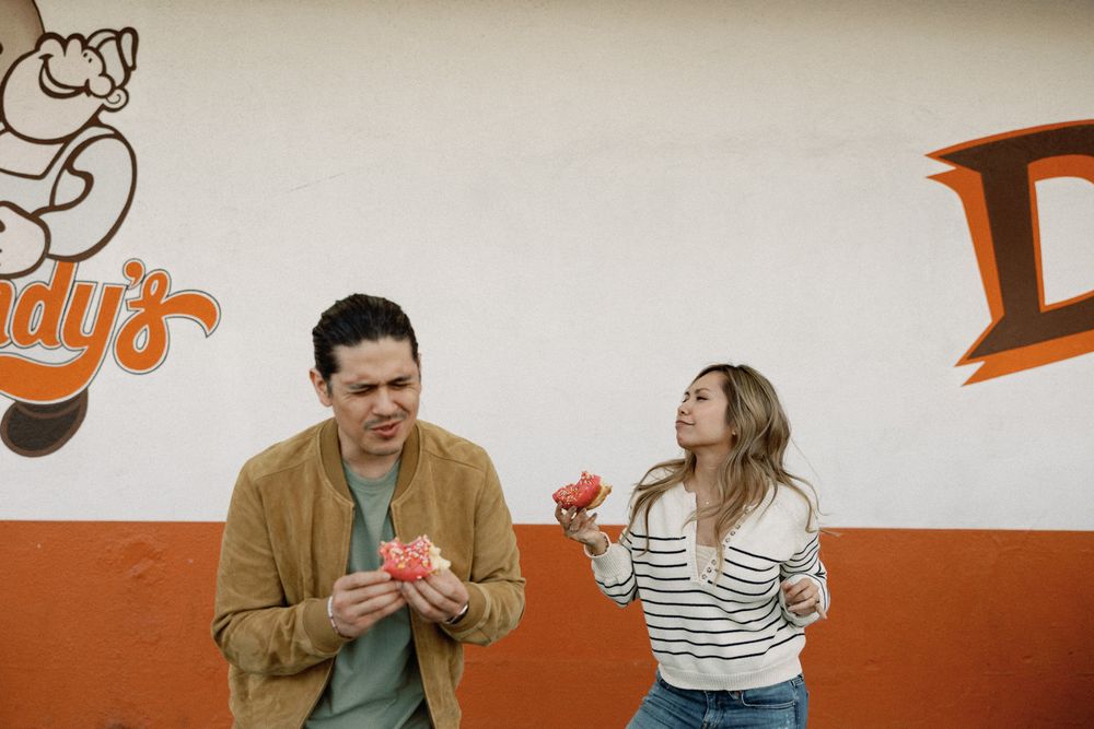 Couple eating food outside a colorful restaurant mural, man in brown jacket and woman in striped shirt enjoying snacks.