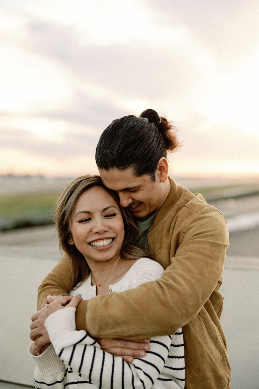 Couple embracing and smiling at sunset, woman in striped sweater, man in brown jacket hugging her from behind.