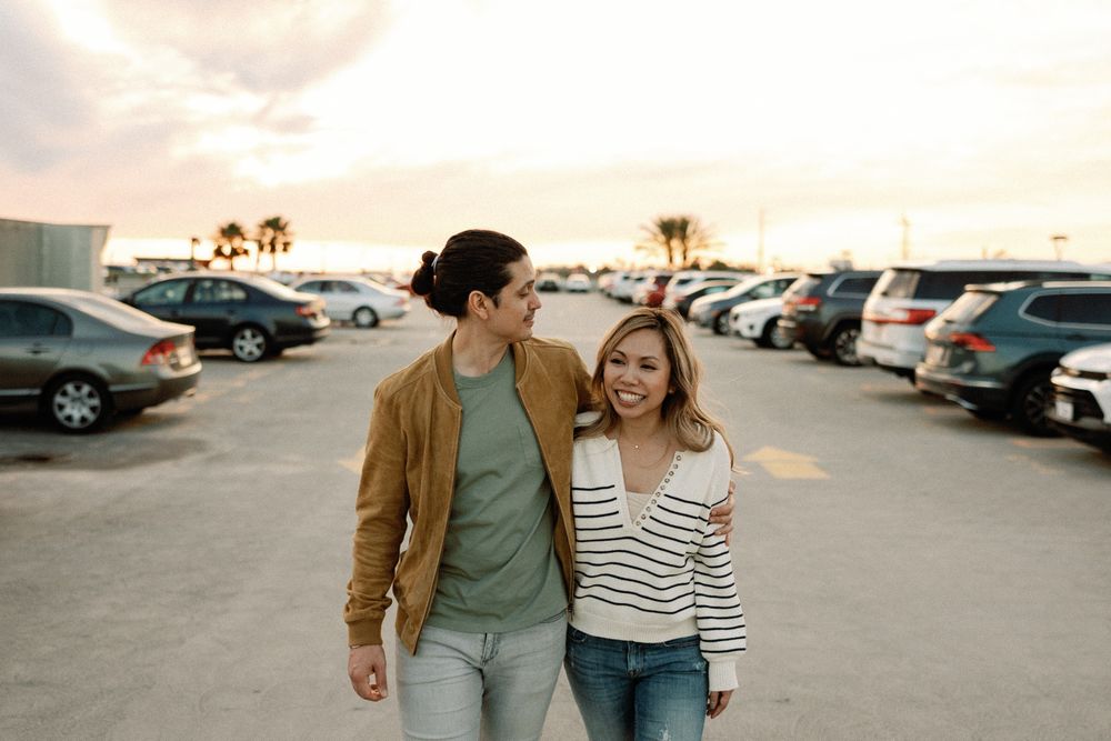 Couple smiling and walking together in a sunny parking lot, man in brown jacket with arm around woman in striped shirt.