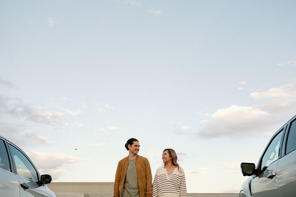 Couple standing between two cars under a blue sky at sunset, smiling and looking at each other.