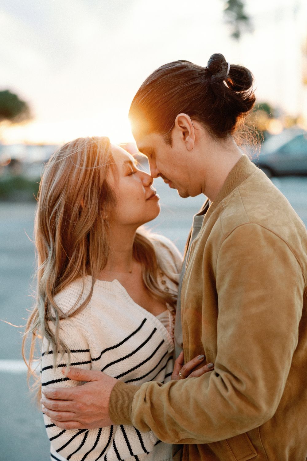 Romantic couple sharing a tender moment at sunset, woman in striped top and man in tan jacket embracing outdoors.