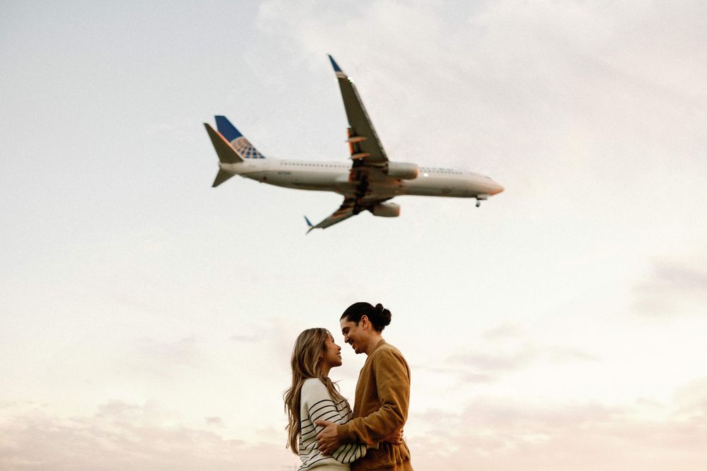 Romantic couple almost kissing with a United Airlines plane flying overhead against a pastel sky.