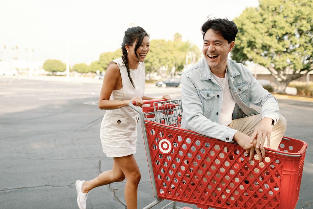 A laughing Asian couple enjoys a fun moment with a red Target shopping cart in a sunny parking lot.