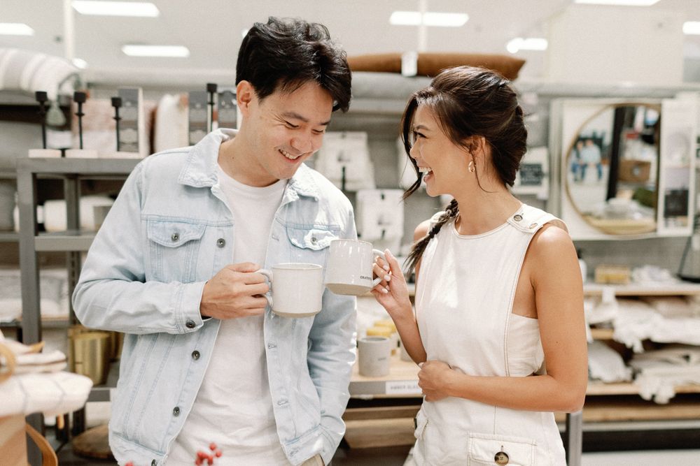 A young Asian couple smiles while browsing mugs in a home goods store, dressed in casual white and denim outfits.