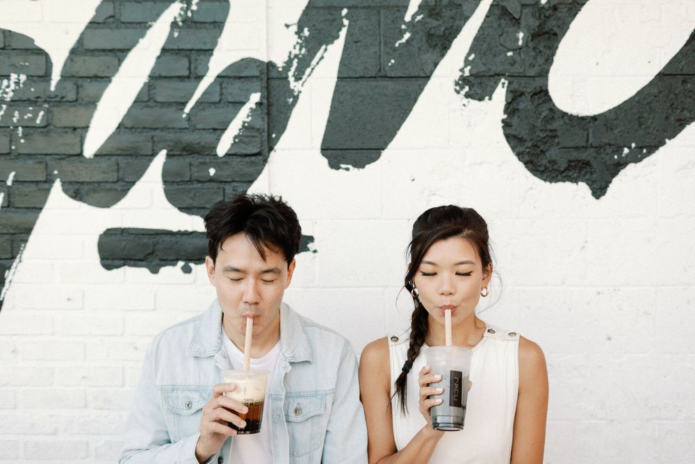 A young Asian couple sipping iced drinks through straws against a white brick wall with black graffiti lettering.
