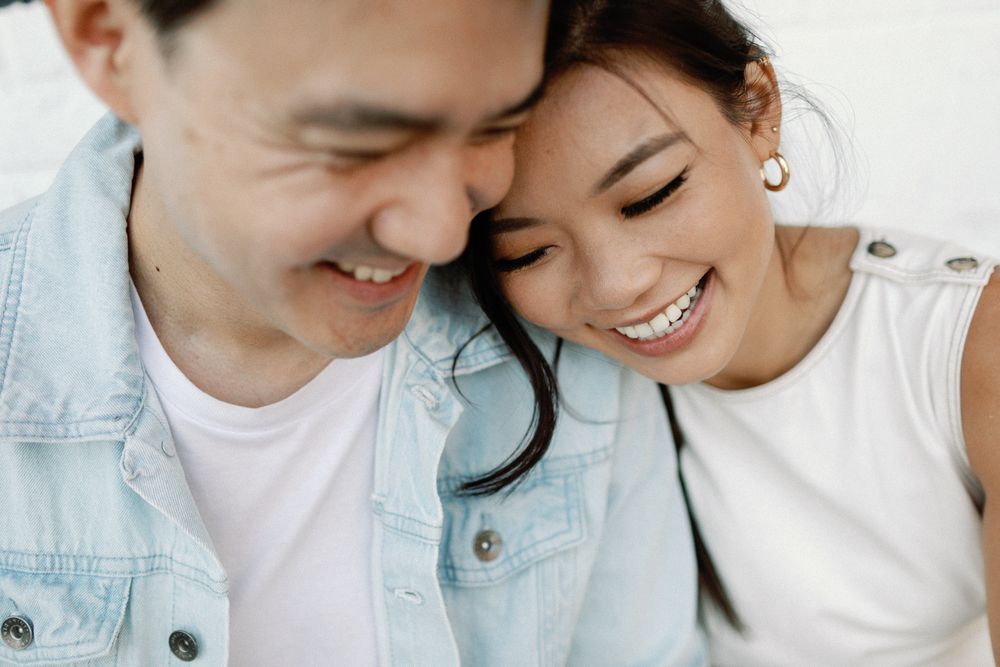 A smiling Asian couple sharing a tender moment, leaning heads together, wearing casual denim and white outfits.