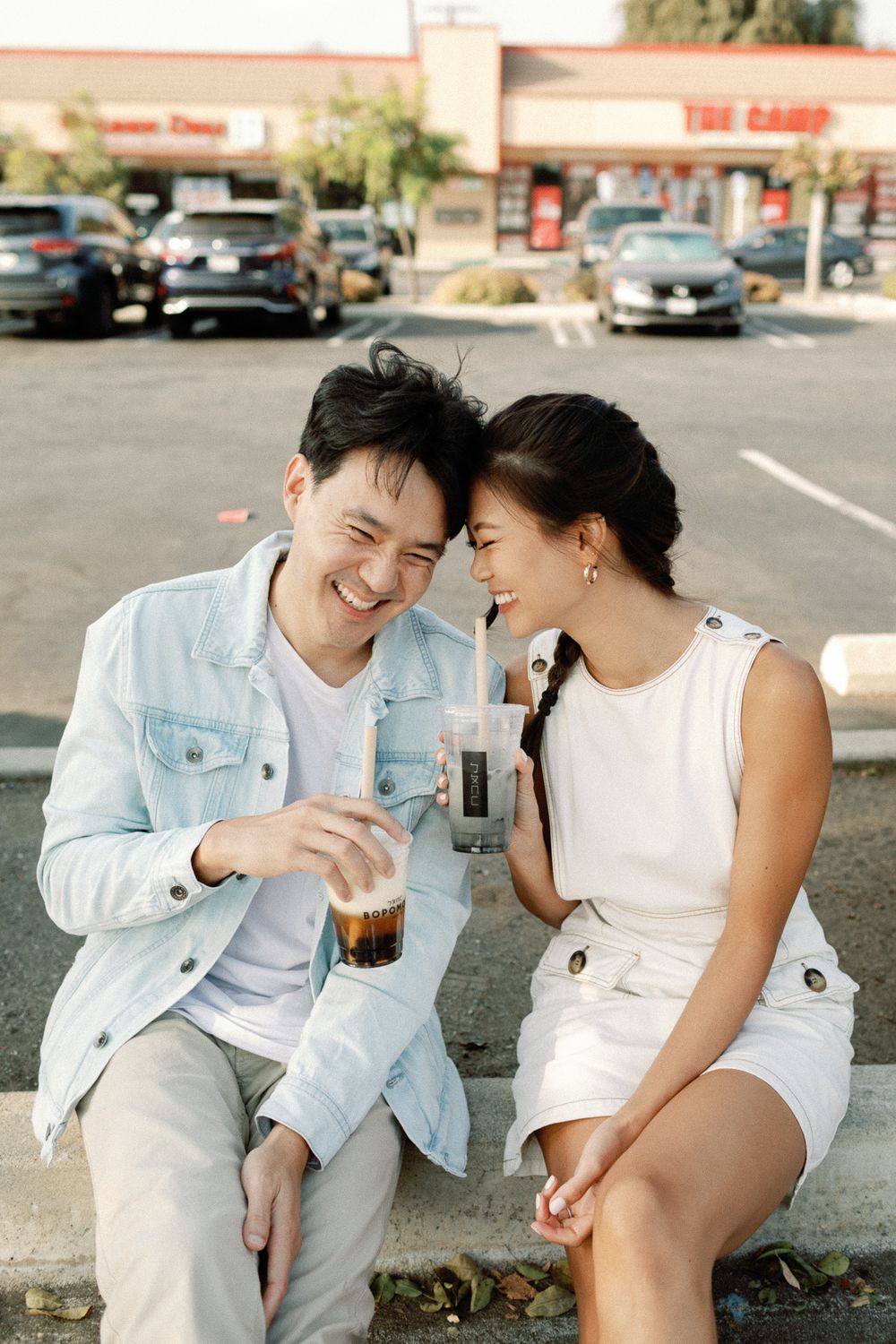 Happy Asian couple sharing iced drinks while sitting together outdoors in a parking lot, smiling and laughing.