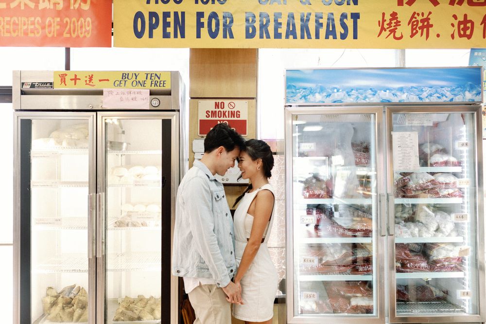 Couple sharing a romantic moment in front of a Chinese bakery open for breakfast, surrounded by refrigerated display cases.