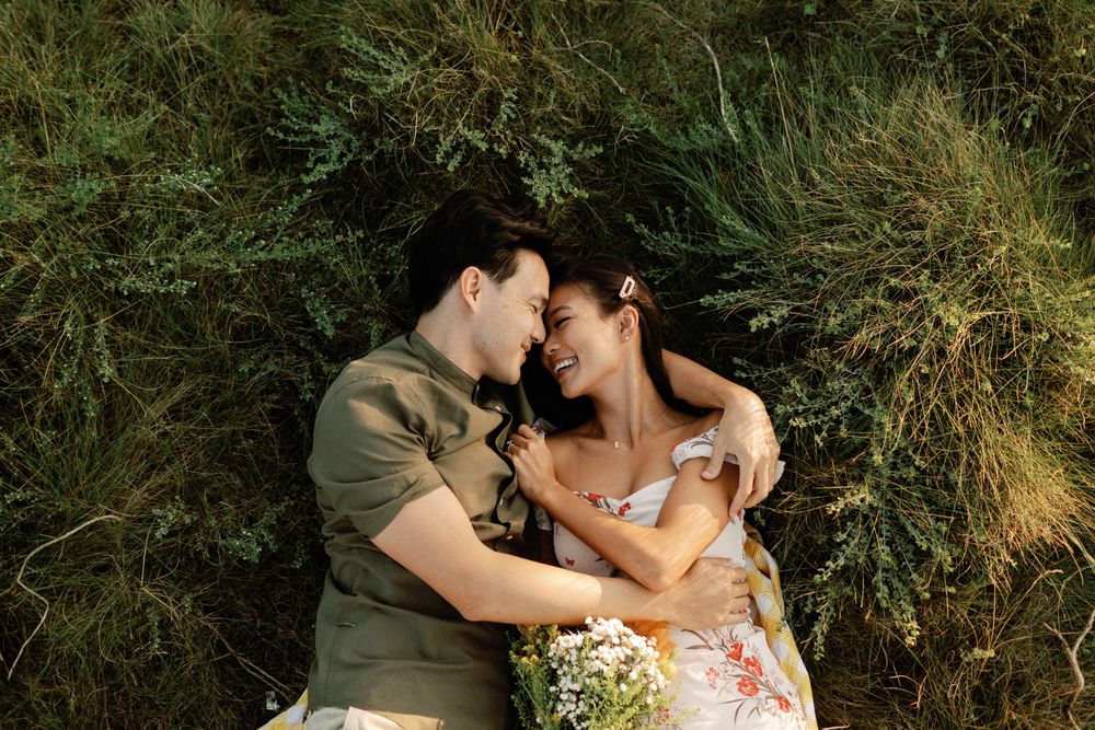 A romantic couple lying together in lush green grass, smiling and gazing at each other tenderly outdoors.