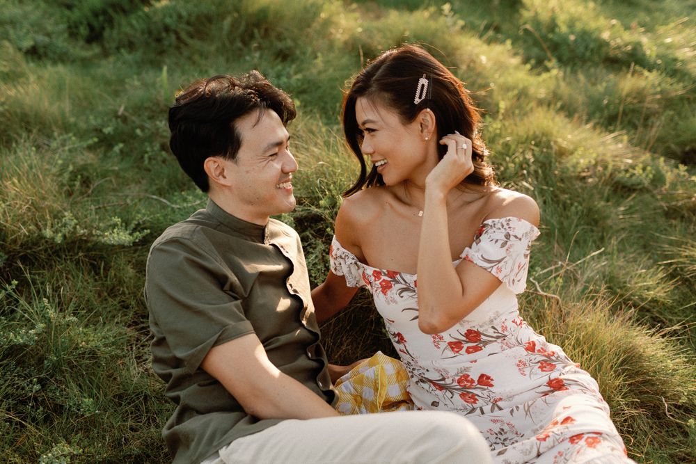 A couple sits together on a grassy hillside, smiling at each other, woman in floral off-shoulder dress.