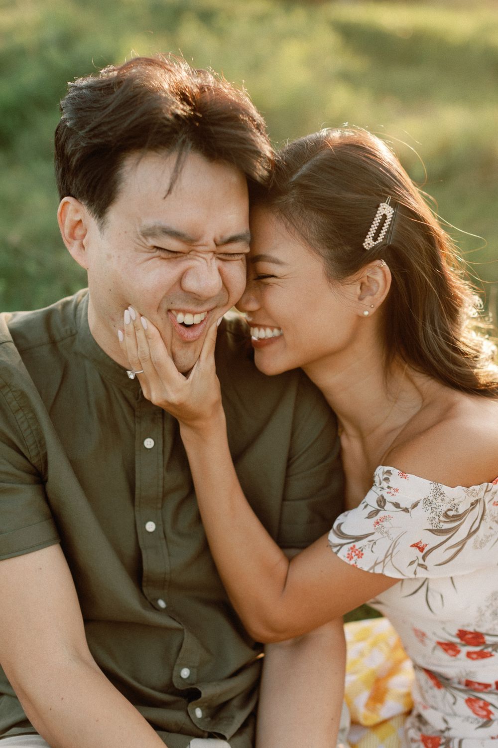 A joyful couple laughing together outdoors, woman in floral dress touching man's face during a sunny picnic.