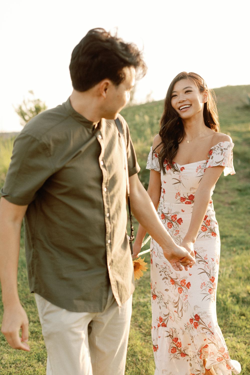 A smiling couple holds hands in a sunny green field, woman in floral off-shoulder dress, man in olive shirt.
