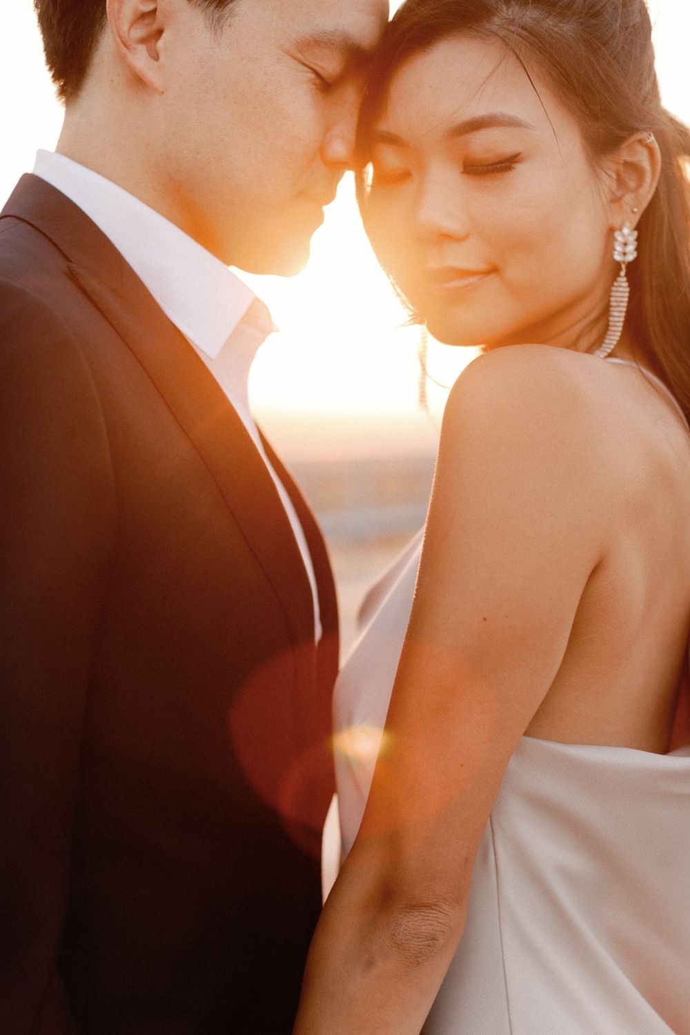 Couple in wedding attire sharing an intimate moment at golden sunset, bride in backless gown with elegant earrings.