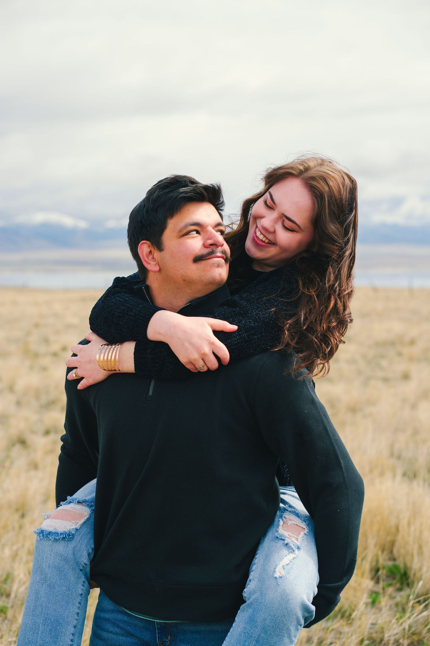 Happy young couple laughing as woman rides piggyback on mans back in golden prairie field.