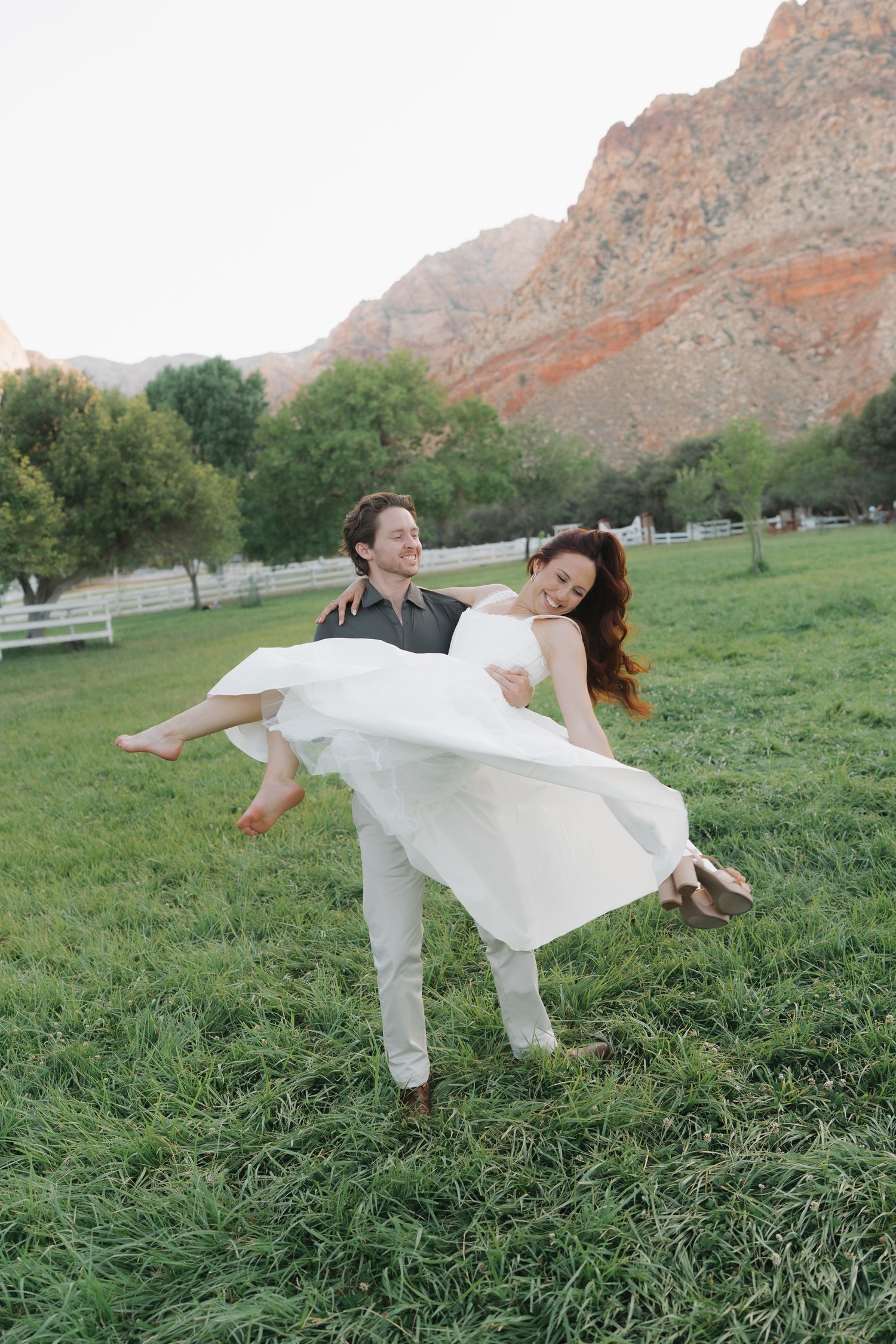 Groom carrying bride in white dress across green meadow with red rock mountains in background.