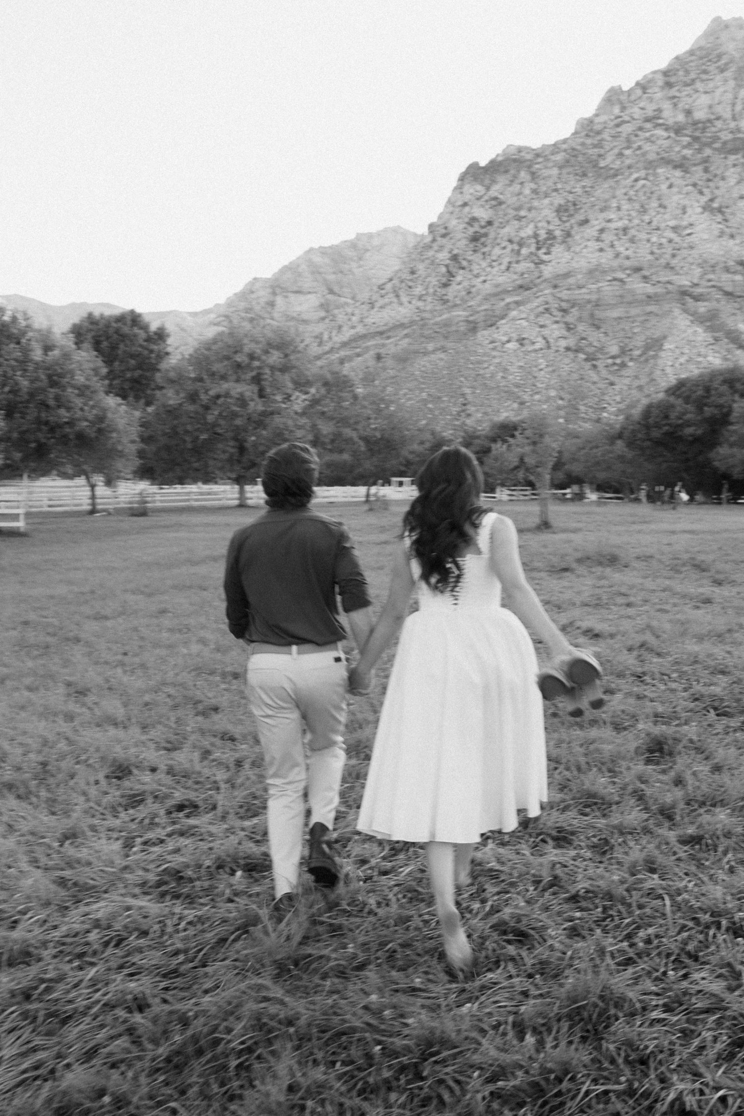 Black and white photo of a couple holding hands walking through a field with mountains in the background.