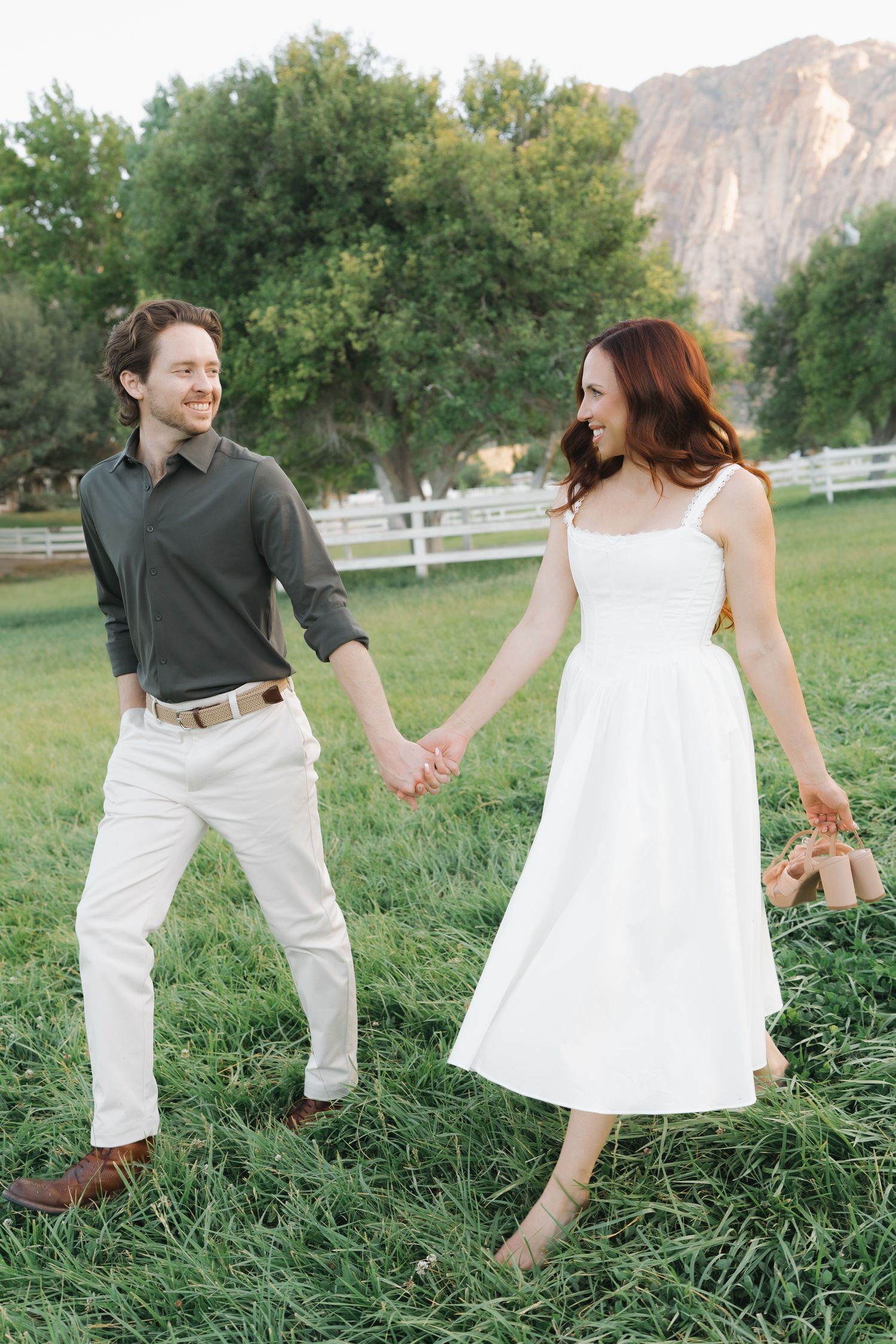 A smiling couple holds hands walking on green grass with white fence and mountain backdrop.