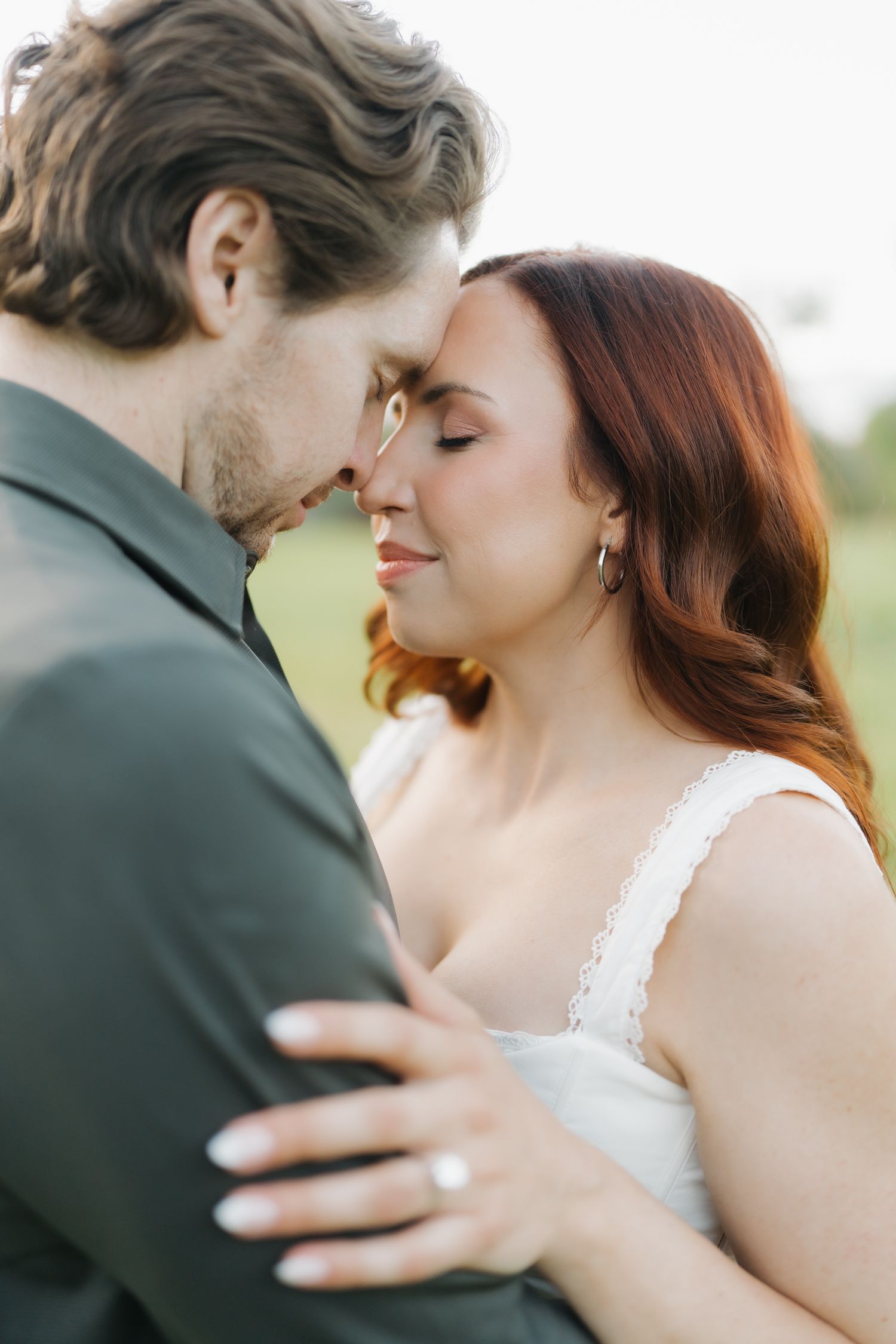 A bride with red hair and groom share an intimate forehead touch moment outdoors in soft natural light.
