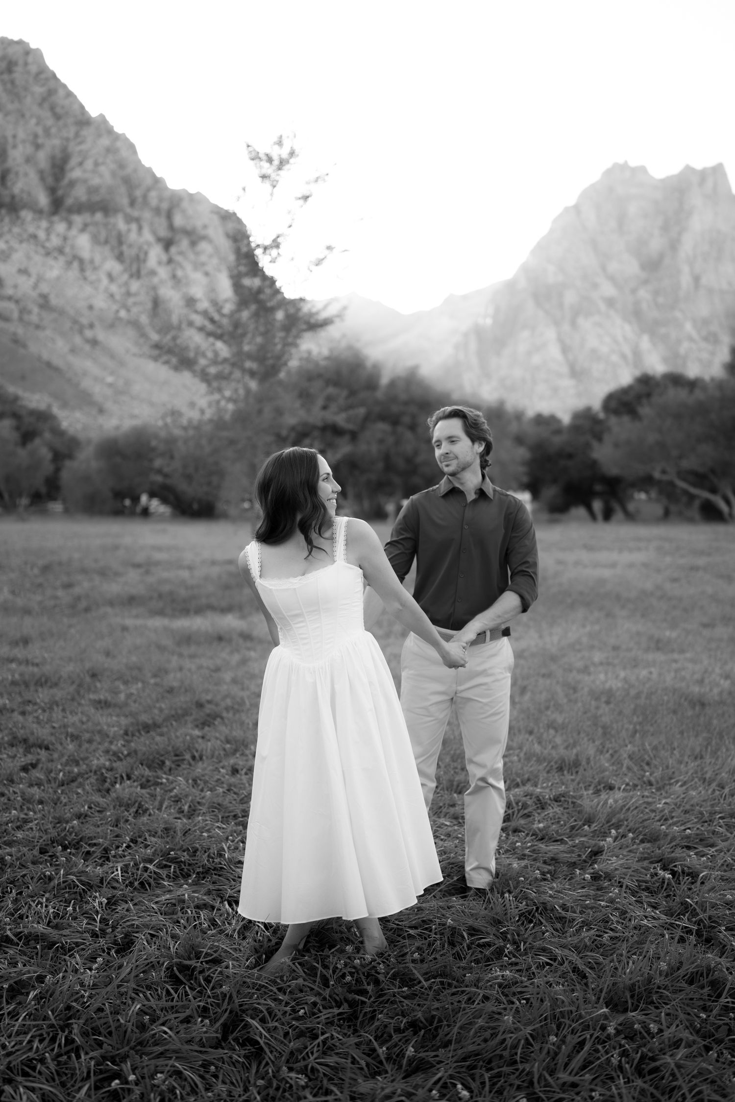 Black and white photo of couple holding hands in field with mountain backdrop at sunset.