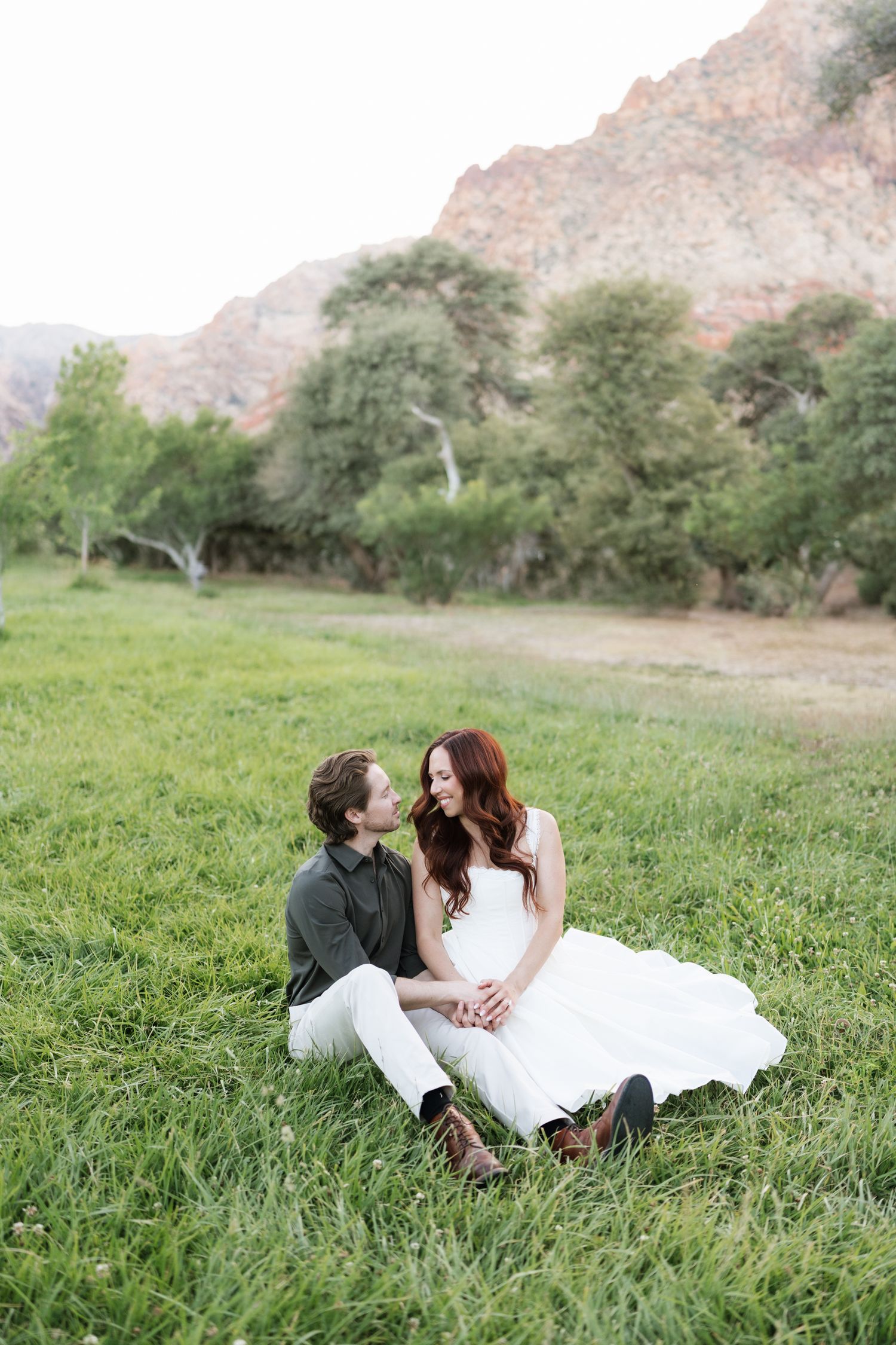 Couple sitting together on lush grass with red rock mountains and trees behind them.