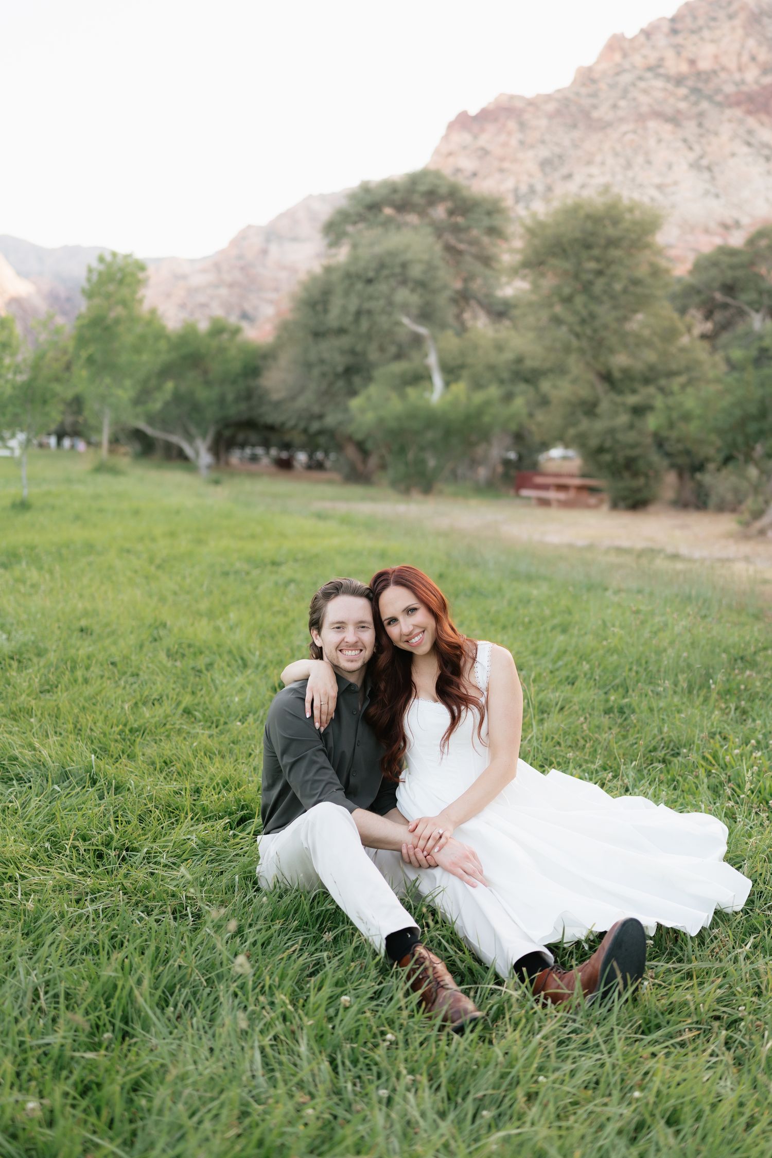 A couple in wedding attire sits together on lush green grass with rocky mountains and trees in the background.