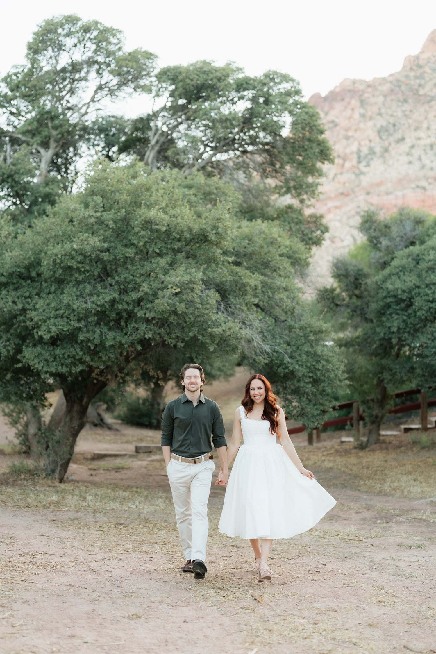 A couple holds hands and walks joyfully through a scenic desert park with lush trees and red rock mountains.