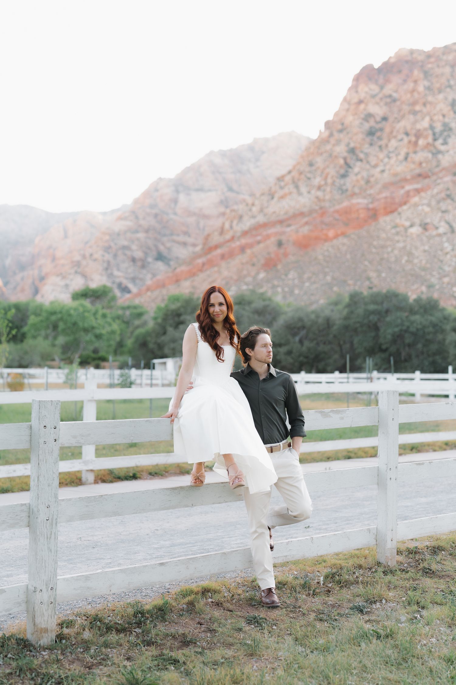 Couple posing by white fence with red rock mountains backdrop, woman in white dress seated on fence rail.