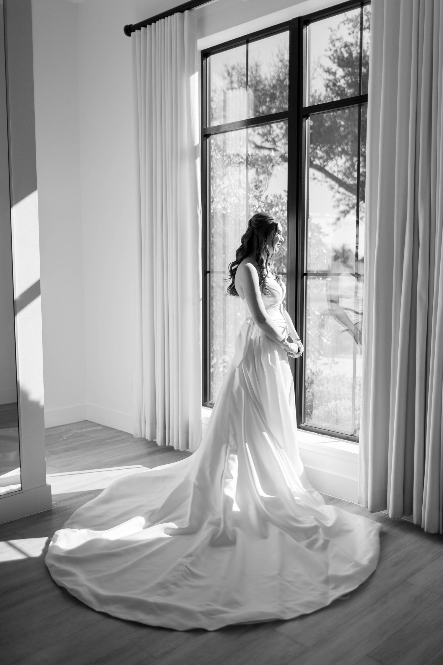 Bride in elegant white wedding gown with long train stands by large black-framed windows in black and white photo.