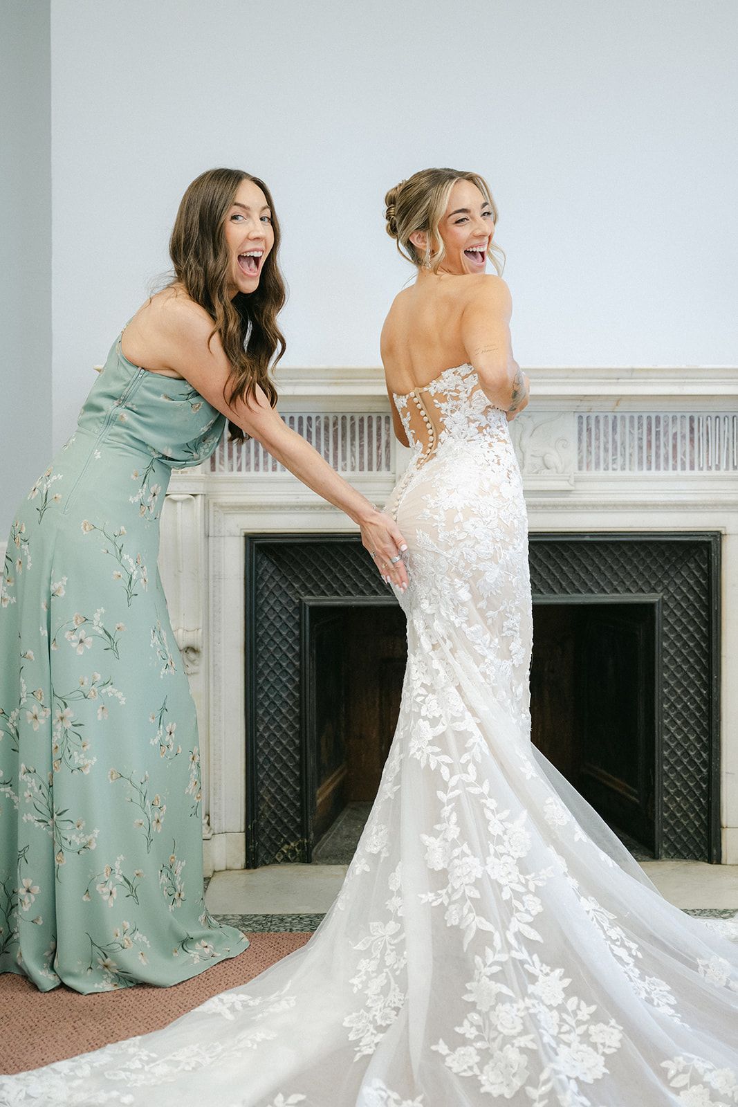 Armour House Wedding - Bride in lace mermaid gown laughing with bridesmaid in sage floral dress near white fireplace.