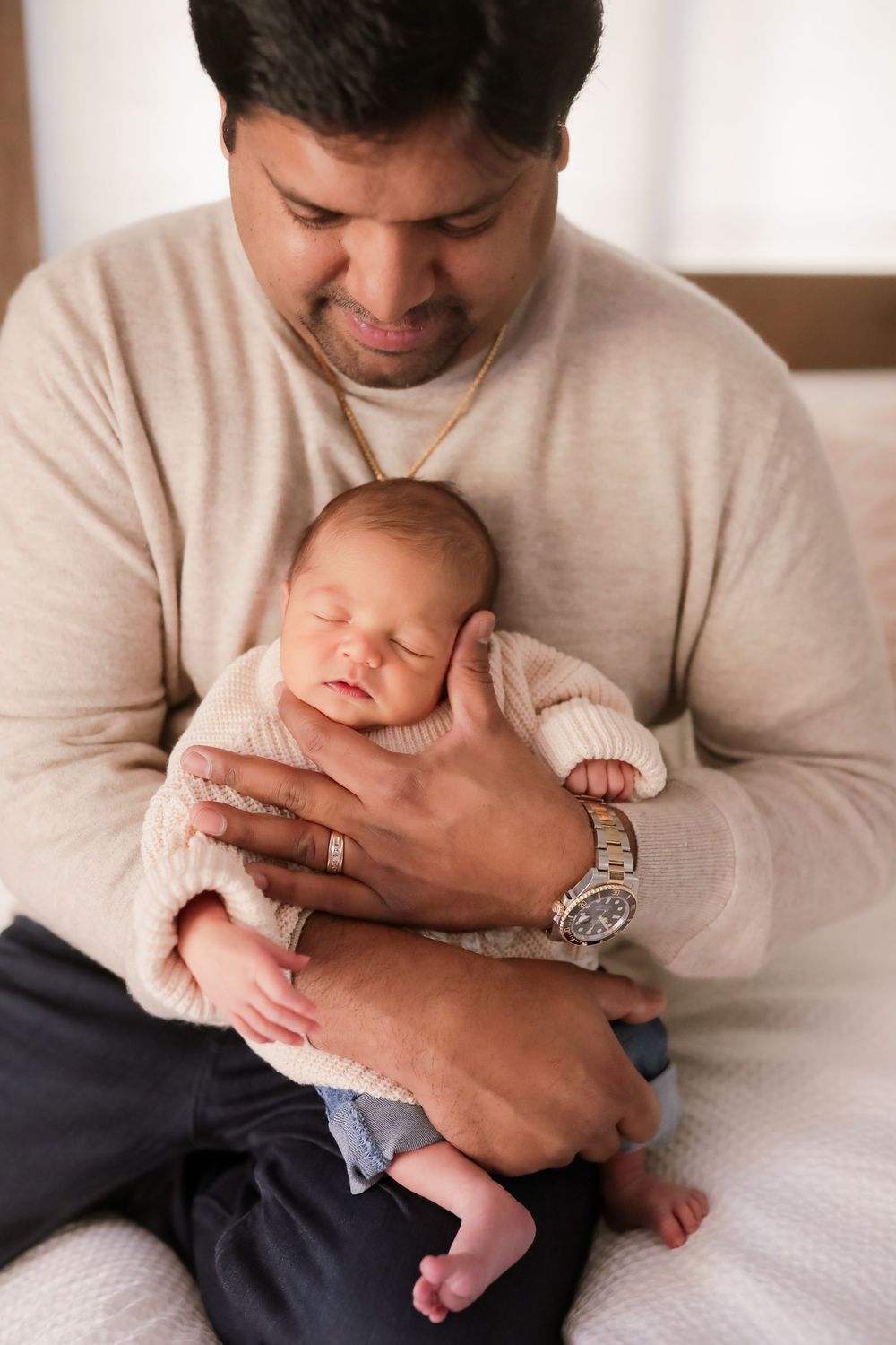 A father tenderly holds his sleeping newborn baby in both hands while sitting on a bed.