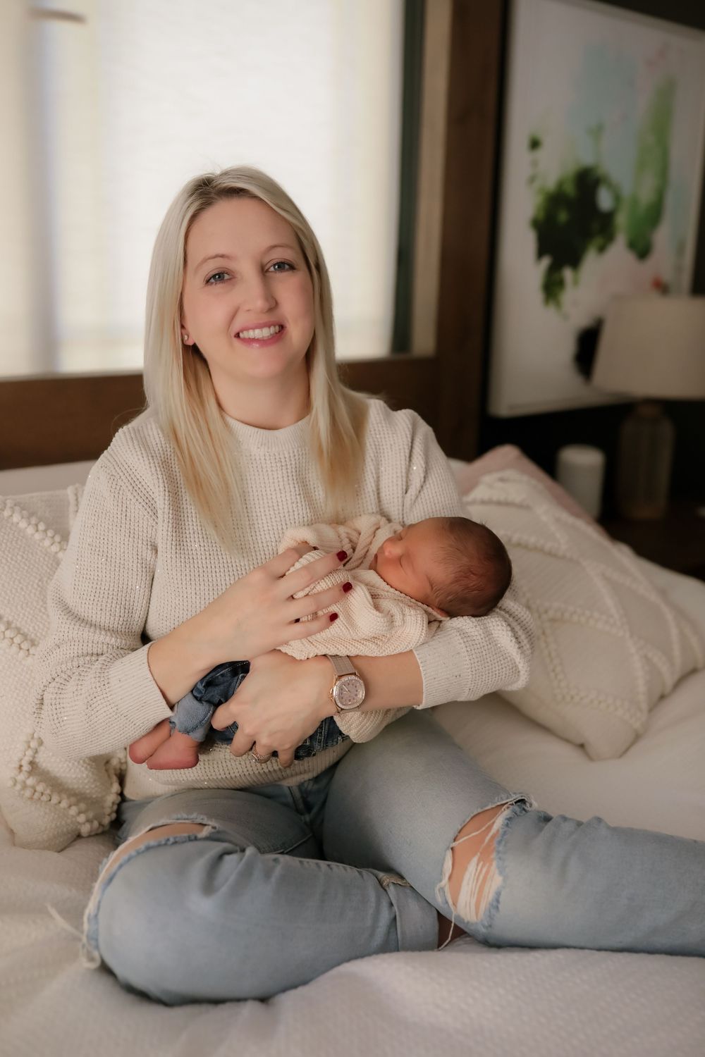 Smiling blonde woman in cream sweater and ripped jeans sitting on bed holding newborn baby in cozy bedroom.