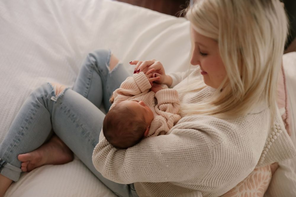 A blonde mother cradles her sleeping newborn baby in her arms while sitting on a white couch.