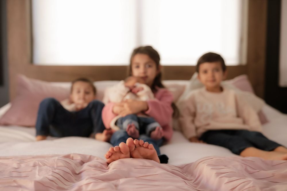 Three children sitting on a bed with pink bedding, with baby feet in the foreground.