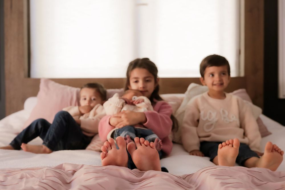 Three young children sitting barefoot on a pink bed, smiling and relaxing together in a cozy bedroom.