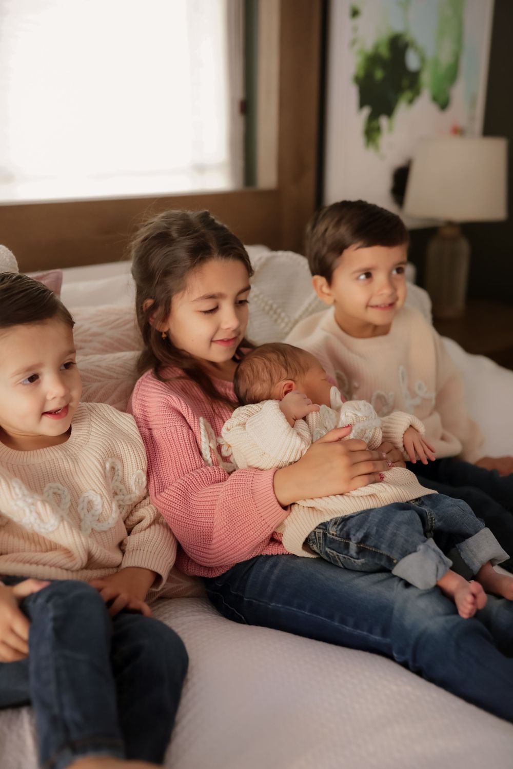 Four siblings sit on a bed, with the oldest girl gently cradling a newborn baby in a warm family moment.