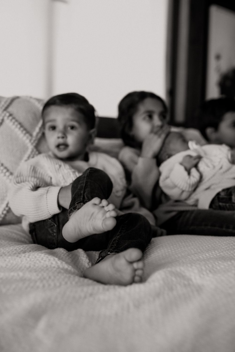 Black and white photo of young siblings sitting on a bed, with a toddler in the foreground showing bare feet.