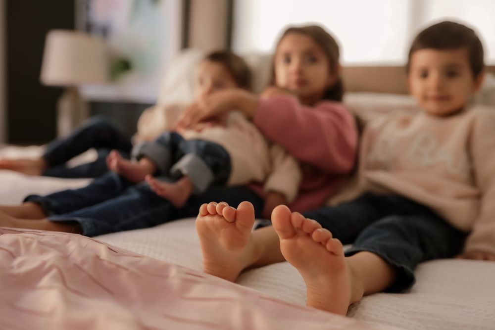 Three young children relaxing on a bed with bare feet in the foreground, captured in a cozy indoor setting.