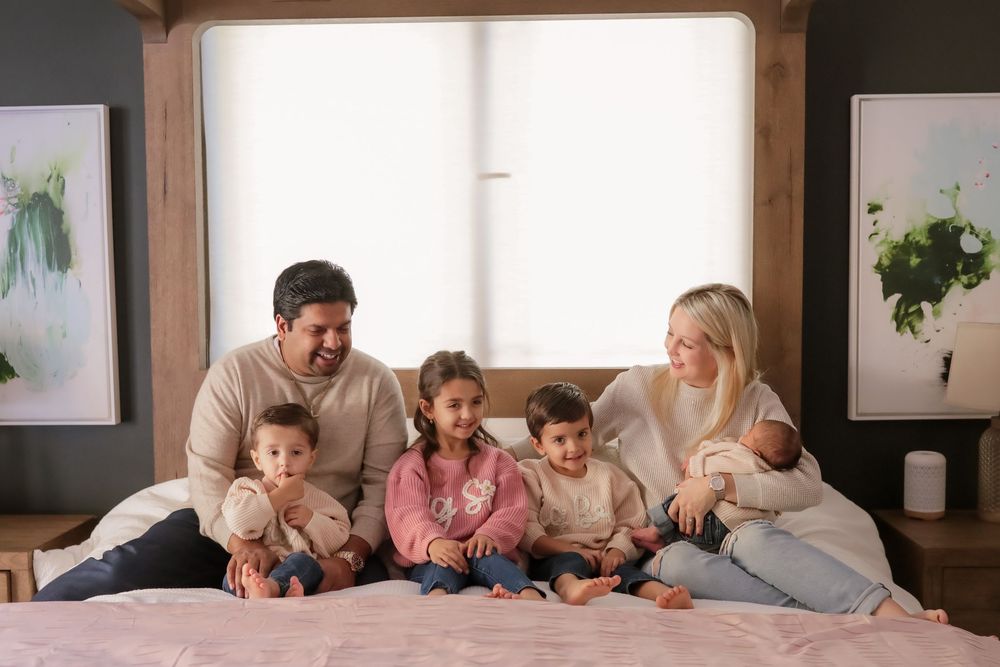 A happy family of six with four children and a newborn sit together smiling on a bed.