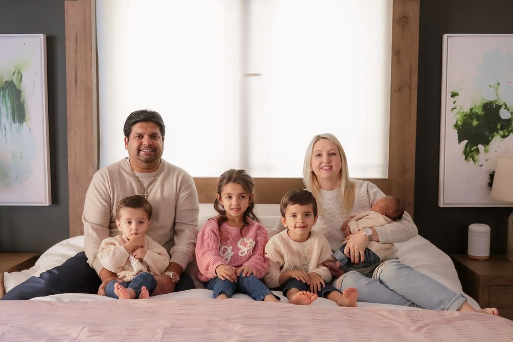 Happy family of six with parents and four young children sitting together on a bed for a portrait photo.
