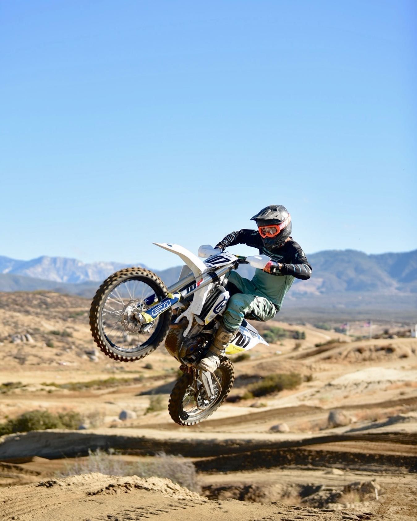 Motocross rider performs a high jump on a dirt bike over a desert track with mountains in the background.