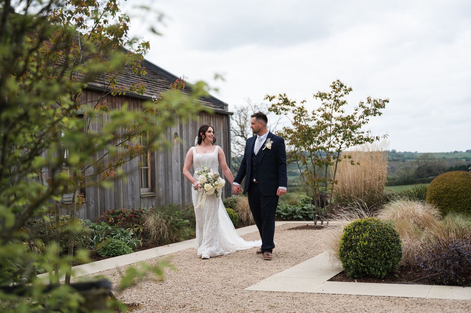 Bride and groom walking hand in hand along a garden path beside a rustic barn venue on their wedding day.