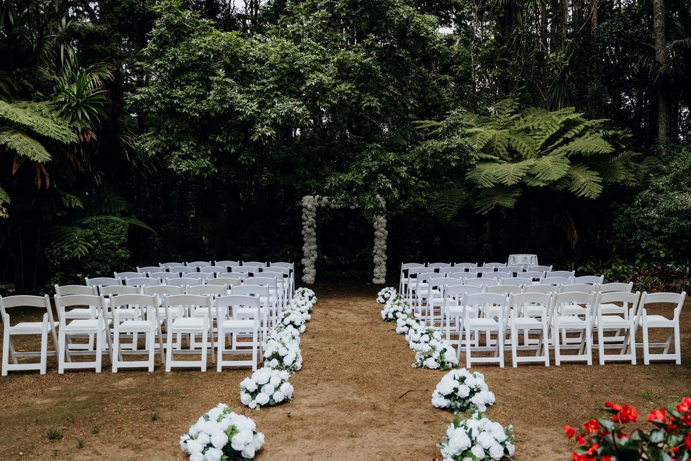 Outdoor wedding ceremony setup with white folding chairs, floral aisle decorations, and a square arch surrounded by lush green trees.